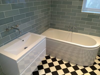 A stylish bathroom with a freestanding bathtub and a modern white vanity unit with integrated sink. The walls are covered in light grey subway tiles, while the black and white checkered floor adds a classic touch. Wall-mounted faucets enhance the sleek, contemporary feel of the space. This bathroom was professionally installed by Martyn Read Plumbing, offering high-quality Norwich plumbing services and Norfolk plumbers.