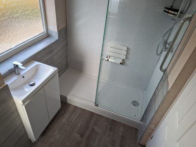 A modern bathroom featuring a walk-in shower with a glass screen and a mounted shower seat. The shower area has light grey walls and a sleek chrome showerhead. Next to the shower is a compact white vanity unit with an integrated sink. The large frosted window allows natural light to fill the space, while the wooden-style flooring adds warmth to the overall design. This bathroom was expertly installed by Martyn Read Plumbing, known for providing Norwich plumbing services and skilled Norfolk plumbers