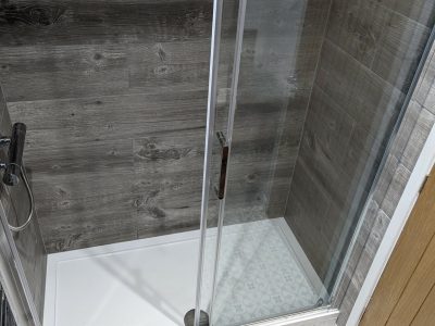 A compact and stylish walk-in shower with sliding glass doors, featuring wood-effect wall tiles and a white shower tray. The floor outside the shower displays intricate patterned tiles, creating a modern and rustic aesthetic. The shower is equipped with sleek chrome fixtures, enhancing its contemporary feel. Installed by Martyn Read Plumbing, this design showcases expert craftsmanship, reflecting the high-quality Norwich plumbing services and Norfolk plumbers provided by the team
