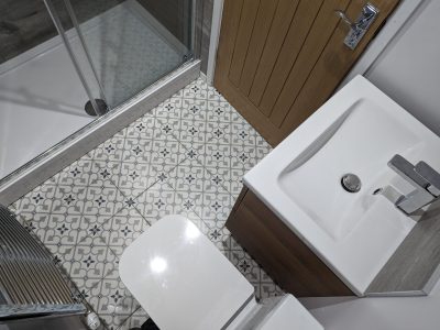A compact bathroom with a white sink, modern faucet, and wooden vanity unit. The walk-in shower with glass sliding doors is positioned next to a toilet, and the floor features beautifully patterned tiles, adding a touch of elegance to the space. A wooden door complements the earthy tones, creating a warm and inviting atmosphere. Expertly designed and installed by Martyn Read Plumbing, specialists in Norwich plumbing services and Norfolk plumbers