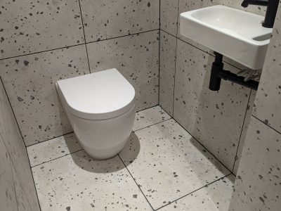A minimalist bathroom with a floating toilet and a small wall-mounted sink, both installed against terrazzo-style tiles with a speckled grey pattern. The modern black fixtures add contrast and a sleek finish to the design. This compact yet stylish bathroom was expertly fitted by Martyn Read Plumbing, providing top-tier Norwich plumbing services and Norfolk plumbers.